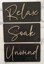 Load image into Gallery viewer, Transform your bathroom into a serene oasis with these Beautiful Relax, Soak, Unwind signs. Crafted from high-quality wood, each sign features delicate embossing of subtle flowers and foliage, creating a soft, nature-inspired background. Perfectly sized at 11 1/2” long x 6” tall, these signs effortlessly add a calming spa-like touch to any space. Whether you're creating a tranquil retreat or enhancing your bathroom's décor, these elegant signs bring a sense of peace and relaxation to your home.