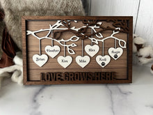 Load image into Gallery viewer, This gorgeous Family Tree Sign is a beautiful and meaningful way to celebrate and honor your family heritage. This personalized frame can be customized with up to 25 names, allowing you to create a unique and special representation of your family. Personalize with up to 25 names, including pets. Hearts hang down from the branches of tree limbs on this beautiful wooden frame. Love Grows Here is displayed across the bottom of the frame.