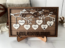 Load image into Gallery viewer, This gorgeous Family Tree Sign is a beautiful and meaningful way to celebrate and honor your family heritage.  This personalized frame can be customized with up to 25 names, allowing you to create a unique and special representation of your family.  Personalize with up to 25 names, including pets.  Hearts hang down from the branches of tree limbs on this beautiful wooden frame.  Love Grows Here is displayed across the bottom of the frame.