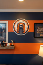 Load image into Gallery viewer, A round decorative sign featuring a stylized Native American chief portrait with a large feathered headdress in navy blue, orange, and white
