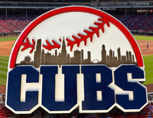 Load image into Gallery viewer, A Chicago Cubs baseball-themed sign or plaque featuring the team name 'CUBS' in large navy letters with white and red outlines, a red baseball seam arc, and a dark city skyline on a white background, set against a blurred green mossy outdoor backdrop.
