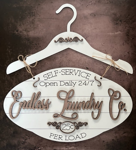 Whimsical Laundry sign. Oval bottom section hangs from a wooden coat hanger with beautiful detail. Sign reads Endless Laundry Co. across the middle in a larger, darker wood lettering. At the top Self Service, Open Daily 24/7 is engraved into the oval, and the bottom reads 25 cents Per Load, which is also engraded into the white background. Makes a beautiful and whimsical addition to any laundry area. Adds a touch of farmhouse feel.