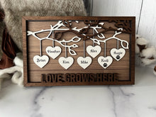 Load image into Gallery viewer, This gorgeous Family Tree Sign is a beautiful and meaningful way to celebrate and honor your family heritage. This personalized frame can be customized with up to 25 names, allowing you to create a unique and special representation of your family. Personalize with up to 25 names, including pets. Hearts hang down from the branches of tree limbs on this beautiful wooden frame. Love Grows Here is displayed across the bottom of the frame.
