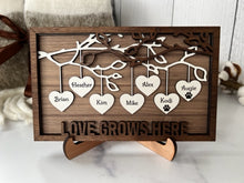 Load image into Gallery viewer, This gorgeous Family Tree Sign is a beautiful and meaningful way to celebrate and honor your family heritage.  This personalized frame can be customized with up to 25 names, allowing you to create a unique and special representation of your family.  Personalize with up to 25 names, including pets.  Hearts hang down from the branches of tree limbs on this beautiful wooden frame.  Love Grows Here is displayed across the bottom of the frame.
